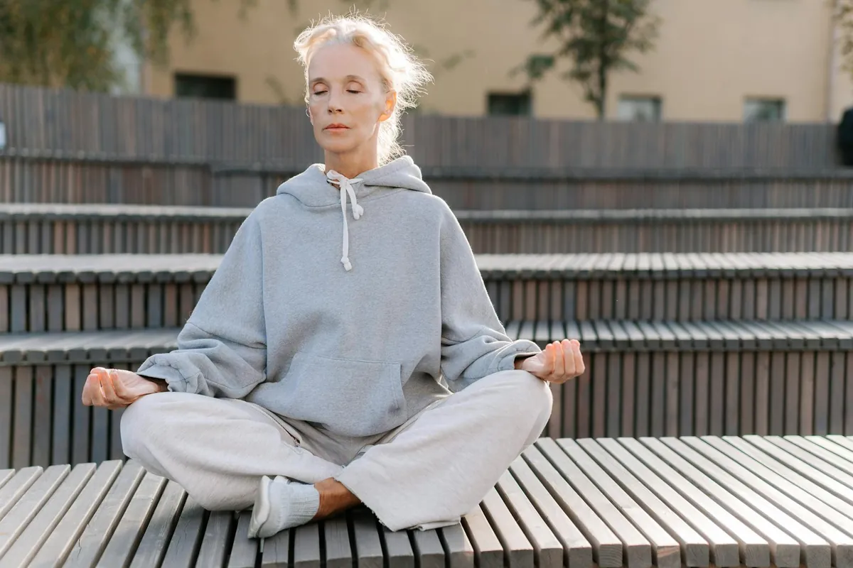 A woman sits calmly with her eyes closed, practicing mindfulness meditation in a park with a blurred city background.