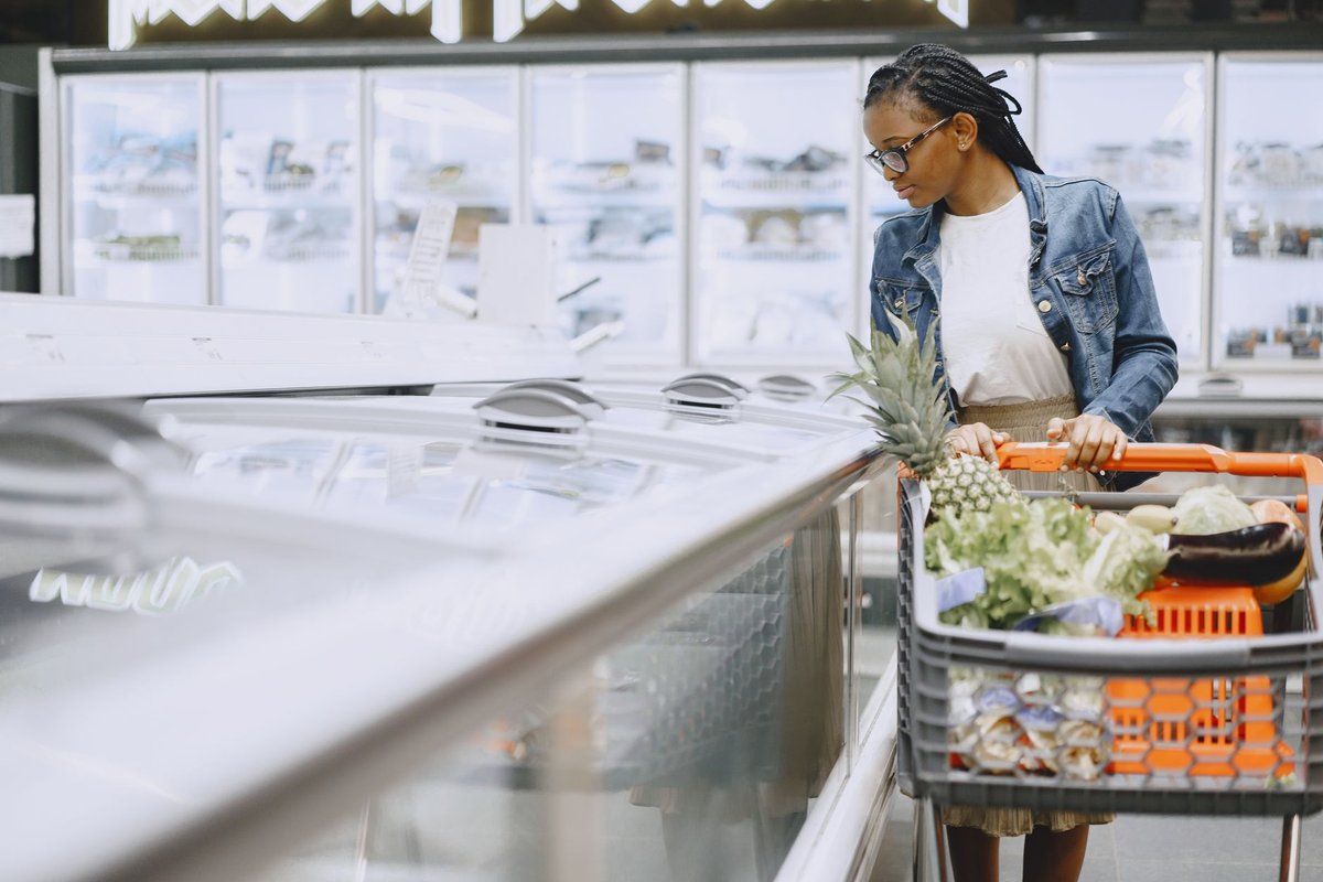 A woman smiling while holding fresh vegetables and packaged groceries in a brightly lit supermarket.